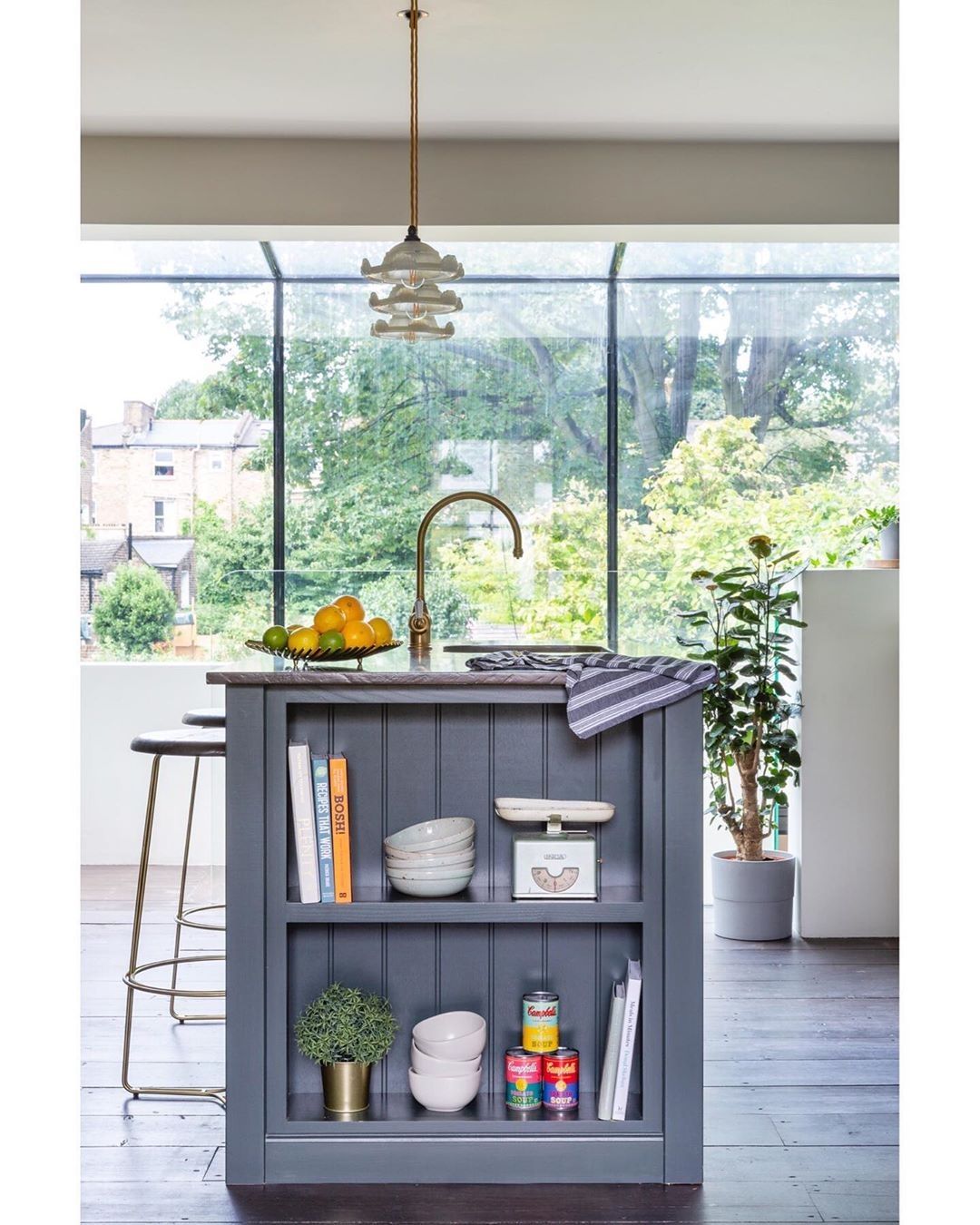 A slim kitchen island styled as a bookshelf, with cookbooks, crockery and potted plants, glass roof and garden visible beyond.