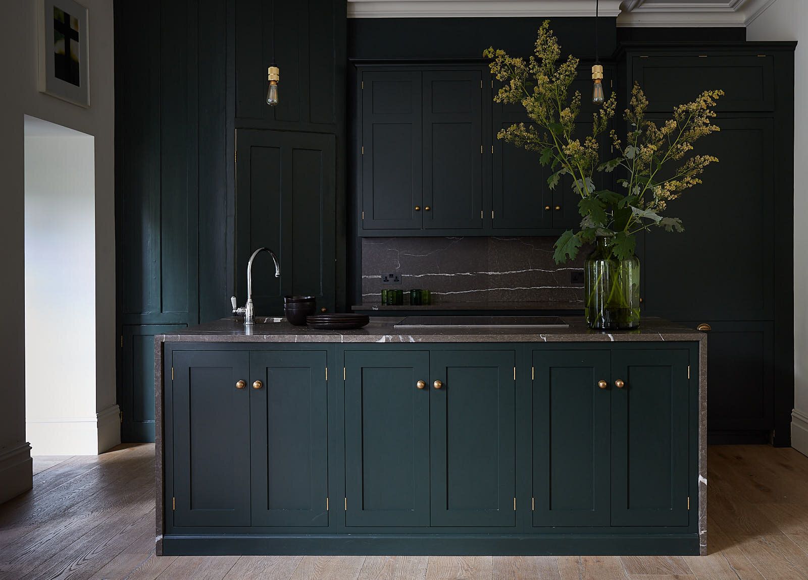 A dark navy shaker-style kitchen with a marble-topped island, brass tap, and a vase of yellow flowers.