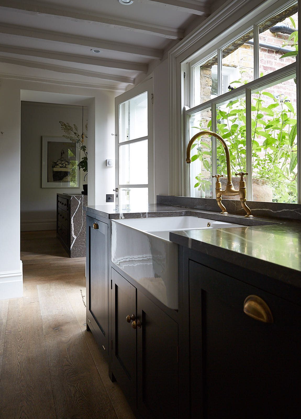 A Belfast sink set into a dark countertop beneath a large sash window, with sunlit greenery visible outside.