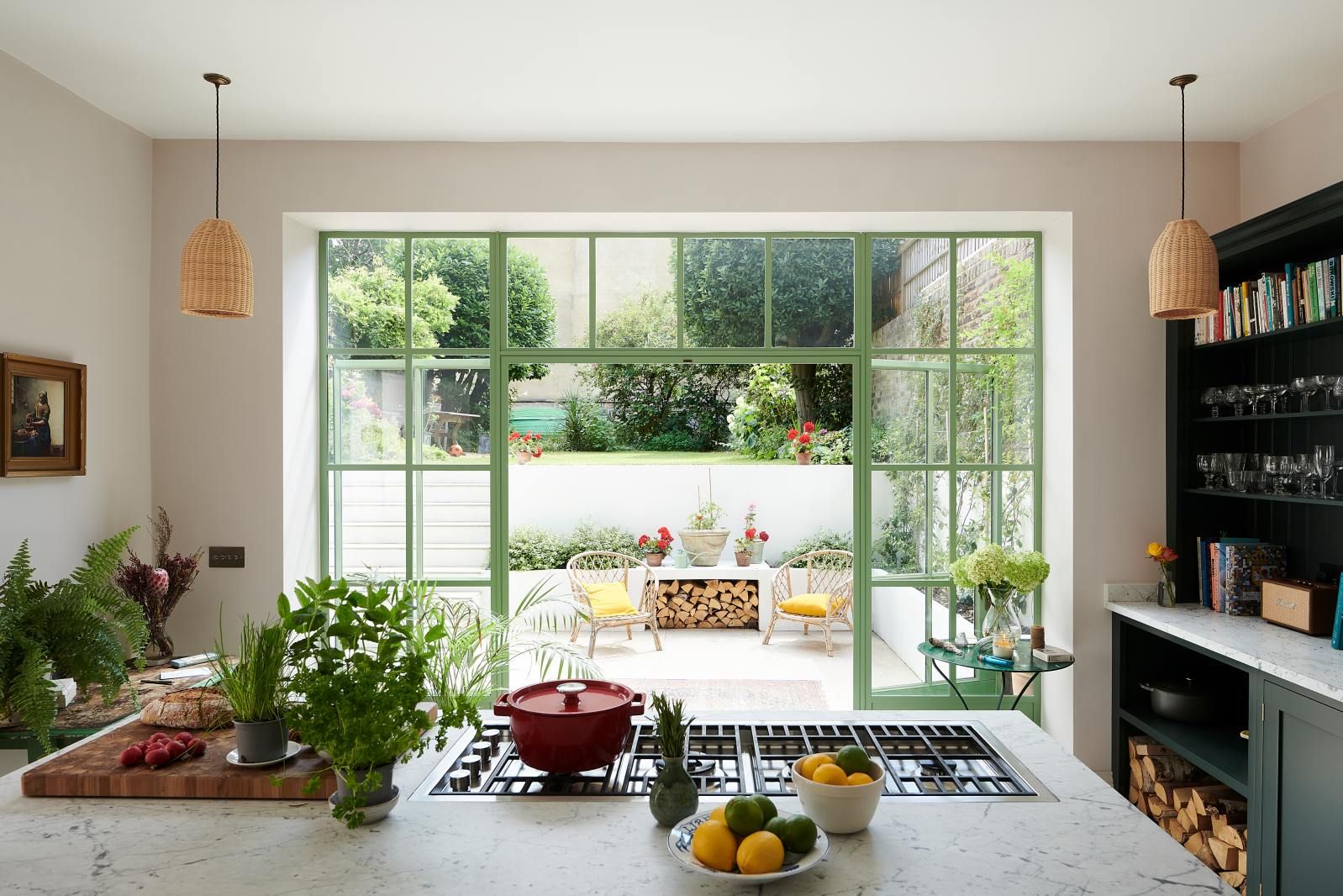 A styled hob scene with a red pan and fresh vegetables and citrus fruit, garden doors open in the background.